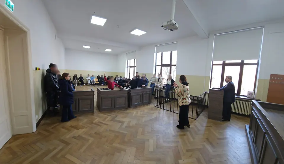 Profesori explicând elevilor organizarea unei săli de judecată, cu boxe din lemn și pupitrul judecătorului
