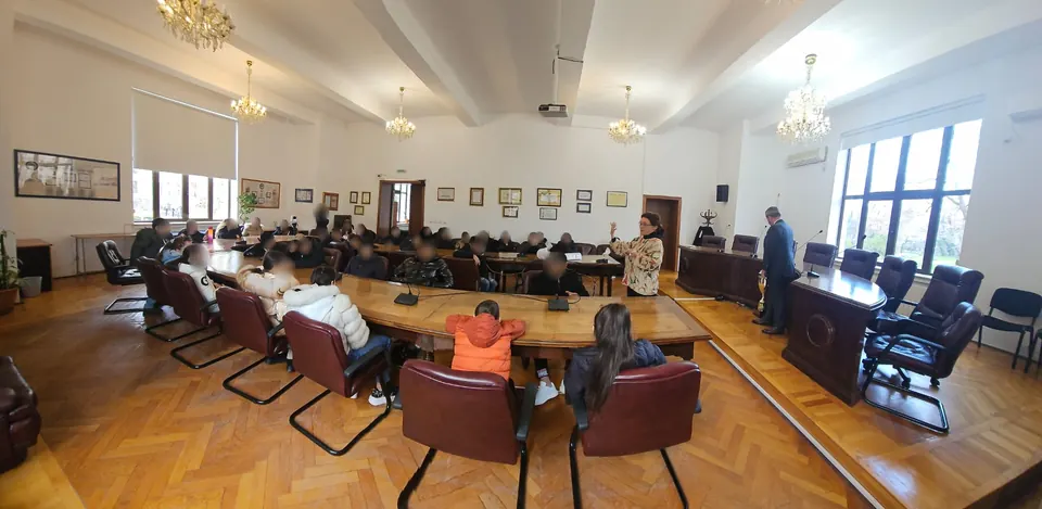 Elevii așezați la masa rotundă din sala de consiliu, în timp ce o profesoară le vorbește