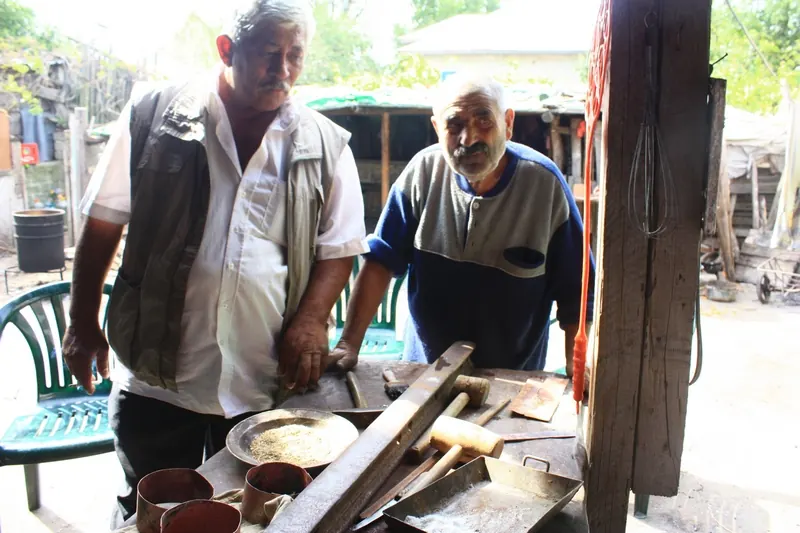 Two Roma coppersmiths working in an open-air workshop, with copper vessels on the table
